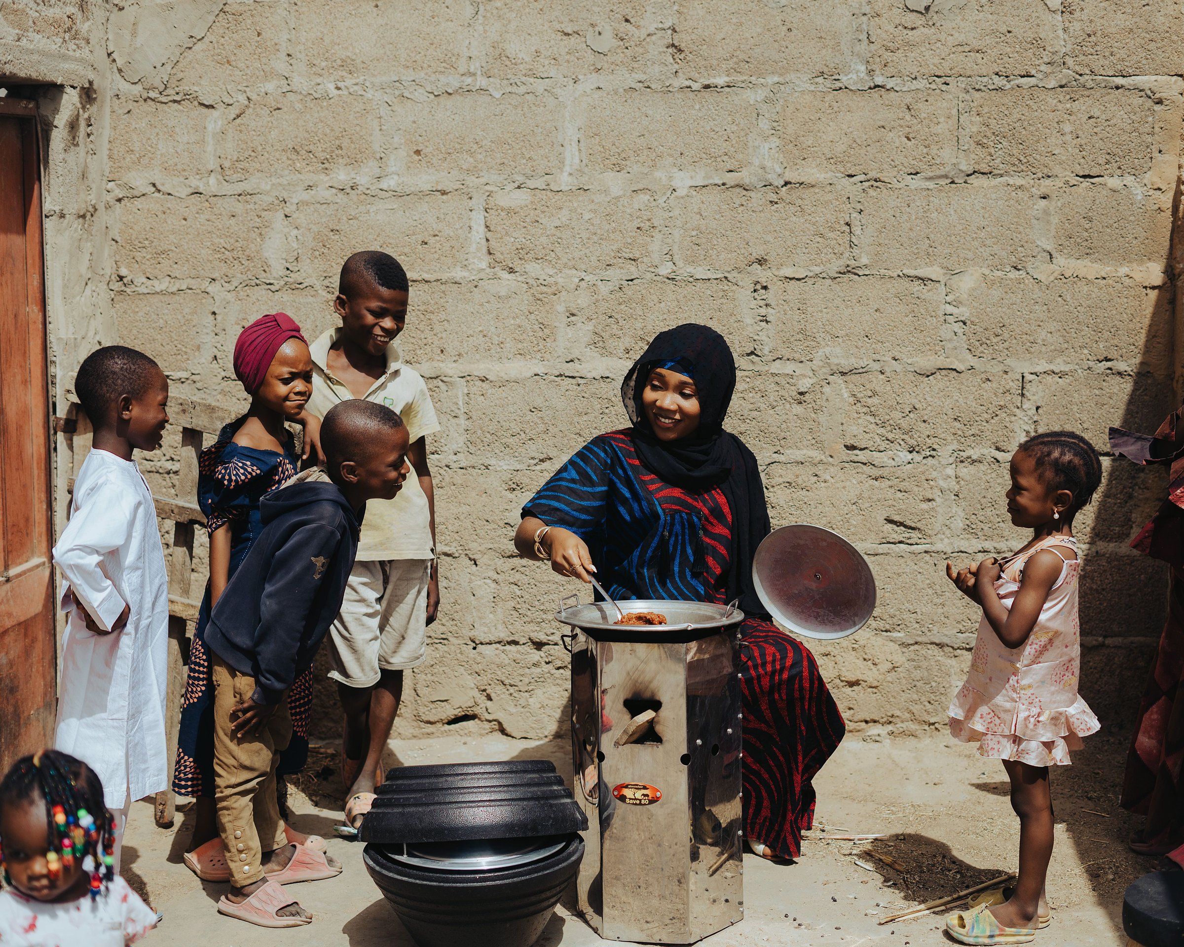 A woman cooks on an efficient stove while children gather around her