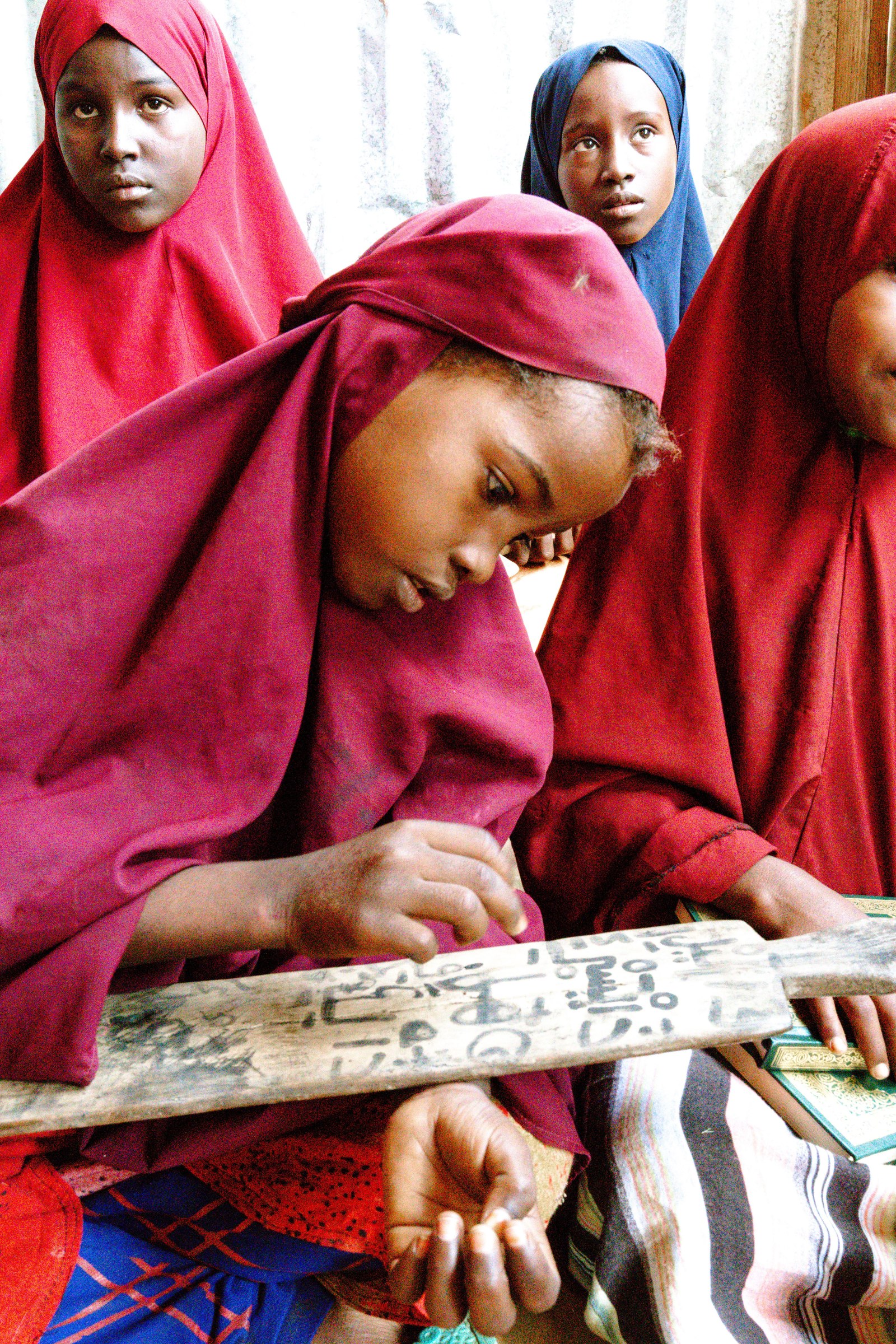 Girls in headscarves write on a wooden slate at a community school